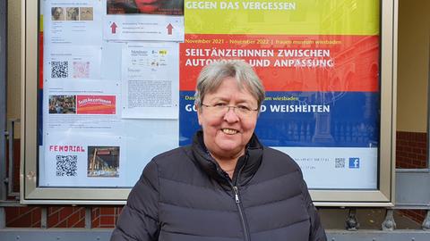 Eine Frau mit kurzen grauen Haaren, rahmenloser Brille und schwarzer Daunenjacke steht vor einem Schaukasten mit bunten Plakaten. Sie lächelt in die Kamera.