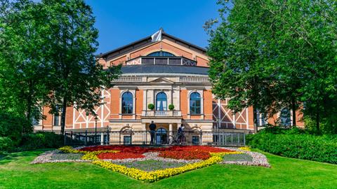 Das Bild zeigt das terracottafarbene Festspielhaus Bayreuth mit einem bunt bepflanzten Blumenbeet davor.