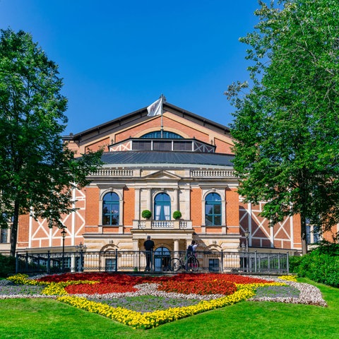 Das Bild zeigt das terracottafarbene Festspielhaus Bayreuth mit einem bunt bepflanzten Blumenbeet davor.