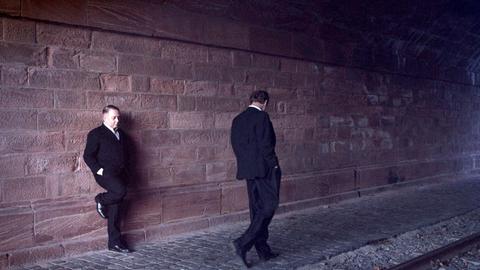 Ein Mann in feinem Anzug lehnt an einer Mauer einer dunklen Unterführung. Ein anderer geht mit gesenktem Kopf vorbei.