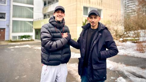 Zwei junge Männer in Daunenjacken vor einem Hochhaus, sie schütteln einander die Hände.