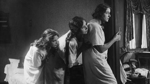 Szene aus dem schwedischen Stummfilm "Norrtullsligan" von 1923. Das Schwarz-Weiß-Bild zeigt drei Frauen in weißen Kleidern, die hintereinander stehen und sich an einer Tür vorbeilehnen, um in einen anderen Raum schauen zu können.