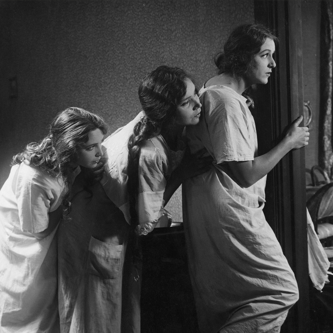 Szene aus dem schwedischen Stummfilm "Norrtullsligan" von 1923. Das Schwarz-Weiß-Bild zeigt drei Frauen in weißen Kleidern, die hintereinander stehen und sich an einer Tür vorbeilehnen, um in einen anderen Raum schauen zu können.