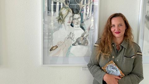 Eine Frau mit dunkelblonden Locken und olivgrüner Jeansjacke steht vor einem Bild zum Grimmschen Märchen  "Brüderchen und Schwesterchen". 