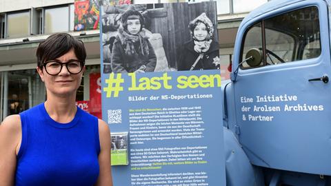 Eine Frau mit Brille steht vor einem alten LKW und einem Banner mit dem Hashtag Last seen