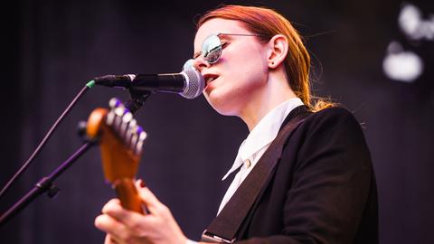 Das Bild zeigt eine Sängerin mit Sonnenbrille, die gleichzeitig Gitarre spielt.
