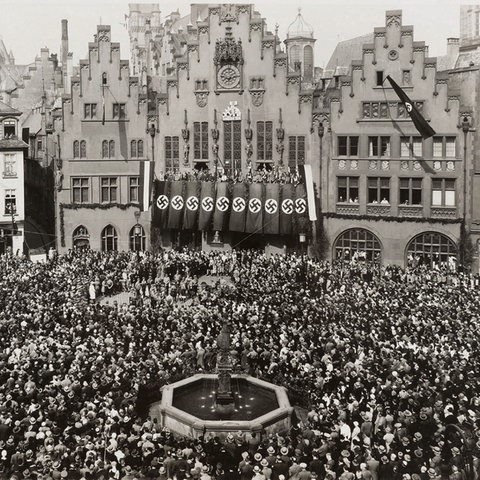Ausstellung "Frankfurt und der NS", Schwarzweiß-Bild des Römerbergs, auf dem sich eine Menschenmasse versammelt hat, am Rathaus hängen Hakenkreuzfahnen