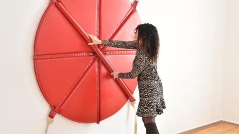 Das Bild zeigt eine Frau mit dunklen Locken und einem schwarz-weiß gemustertem Kleid, die ein an einer weißen Wand montiertes Kunstwerk anfasst. Das Werk besteht aus einem roten lerdenem Kreis, in den vier Einkerbungen eingelassen sind. Ein ebenfalls roter Stab kann in die sich überkreuzenden Einkerbungen gedrückt werden.
