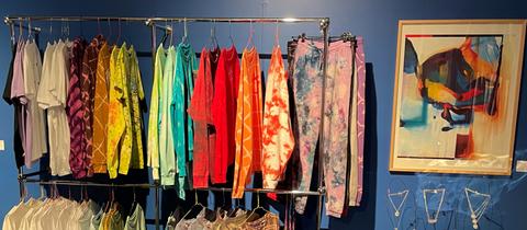 Blick in den Kunst-Pop-Up-Store der Freitagsküche. An einer Kleiderstange hängen Shirts, Hemden und Hosen. An der blauen Wand ein Gemälde, kleine Skulpturen auf einem knallgelben Beistelltisch.