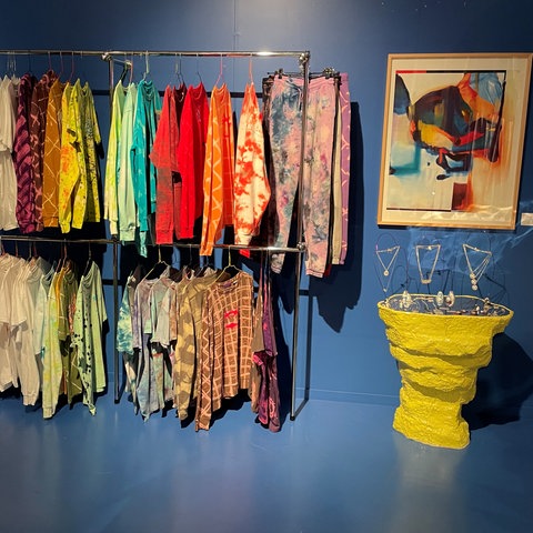 Blick in den Kunst-Pop-Up-Store der Freitagsküche. An einer Kleiderstange hängen Shirts, Hemden und Hosen. An der blauen Wand ein Gemälde, kleine Skulpturen auf einem knallgelben Beistelltisch.