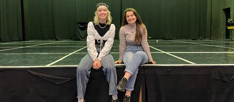 Zwei junge Mädchen sitzen am Rand einer Bühne, die Beine baumeln von der Kante