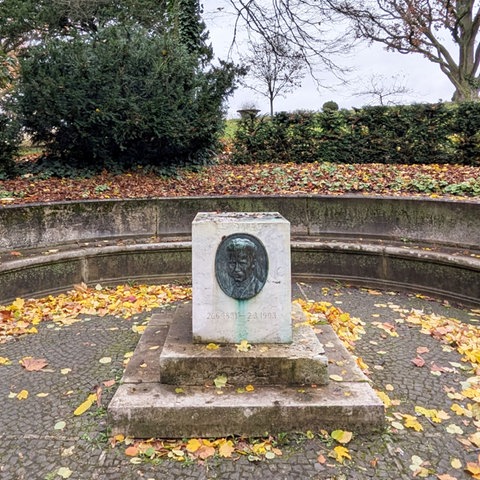 Leerer Sockel auf einem Platz, dahinter eine Hecke, Bäume und rechts ein Gebäude mit teils goldenem Dach.