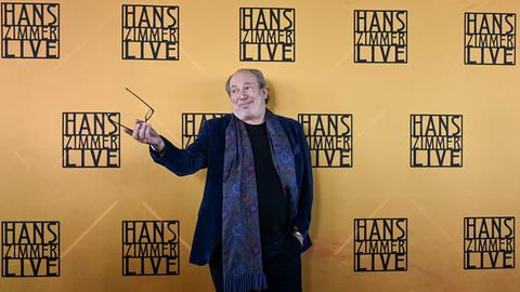 Mann gestikuliert mit der Brille vor einer Wand mit dem Schriftzug "Hans Zimmer live"