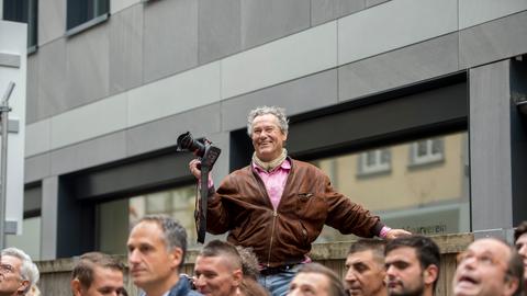 Helmut Fricke steht leicht erhöht in einer Menschenmasse. Er lacht und hat den Arm gehoben, in der Hand seine Kamera.