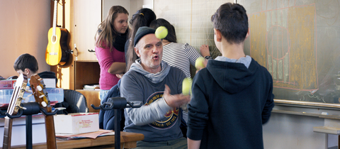 Herr Bachmann im Klassenzimmer, mit Tennisbällen jonglierend und umgeben von Schülerinnen und Schülern.