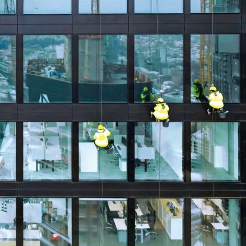 Drei Menschen in gelben Schutzjacken hängen an Seilen an der Glasfassade eines Hochhauses, im Hintergrund die Stadt.