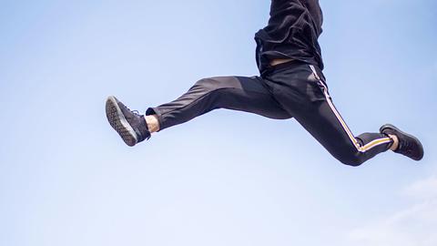 Ein Mann mit Jogginghose springt - vor blauem Himmel aus der Froschperspektive fotografiert.