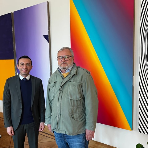 Das Bild zeigt zwei Männer vor einer Wand, an der großformatige bunte Bilder auf Leinwand hängen. Der Frankfurter Oberbürgermeister Mike Josef steht links und trägt einen grauen Anzug. Rechts davon steht der Frankfurter Künstler Tobias Rehberger. Er trägt eine grüne Windjacke und Jeans.