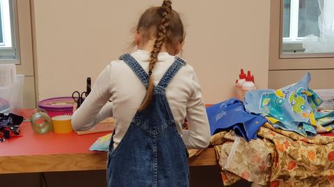 Ein Mädchen mit einem langeb Zopf steht an einem Tisch mit bunten Stoffen, von hinten fotografiert.