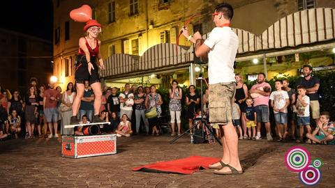 Das Bild zeigt eine Veranstaltung des Straßentheaterfests in Darmstadt. Eine Frau mit einem herzförmigen Luftballon auf dem Kopf steht auf einer Kiste, ihr gegenüber steht ein Mann mit Pfeil und Bogen, der auf den Luftballon zielt. Dahinter sind Zuschauer zu sehen.