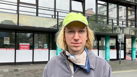 Mann mit langen blonden Haaren und Basecap steht vor einem Haus mit verspiegelter Fassade