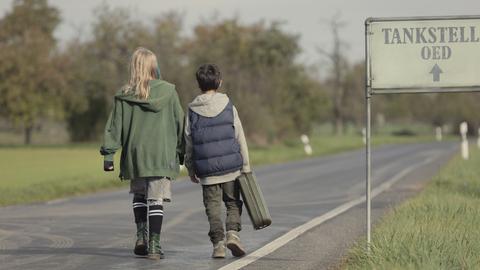 Szene aus dem Kinderfilm "Kannawoniwasein". Ein Junge und ein Mädchen laufen auf einer geteerten Straße, rundherum sind grüne Wiesen. Die Kinder sind von hinten zu sehen, der Junge hat einen Koffer in der Hand.