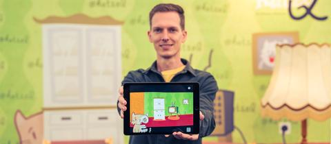 Ein Mann hält ein Tablet nach vorne. Darauf ist die Zeichnung einer Katze zu sehen, die in einem Wohnzimmer vor einem Fernseher sitzt.