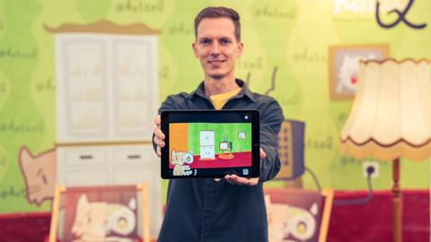 Ein Mann hält ein Tablet nach vorne. Darauf ist die Zeichnung einer Katze zu sehen, die in einem Wohnzimmer vor einem Fernseher sitzt.