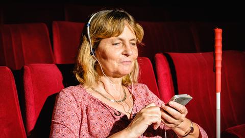 Eine blinde Frau sitzt mit ihrem Stock und ihrem Handy mit einem Kopfhörer auf im Zuschauerraum im Theater.