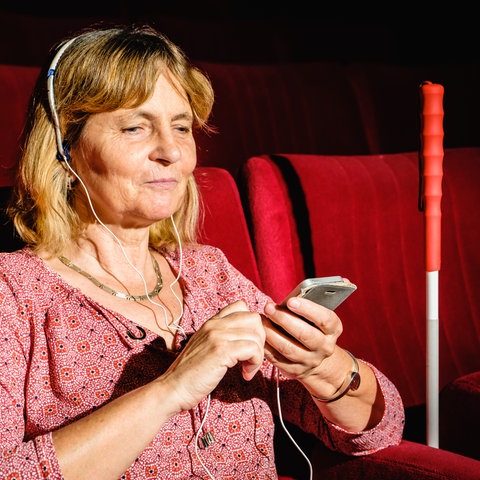 Eine blinde Frau sitzt mit ihrem Stock und ihrem Handy mit einem Kopfhörer auf im Zuschauerraum im Theater.