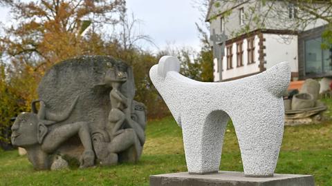 Zwei steinerne Skulpturen in einem Garten, im Hintergrund ein Haus.