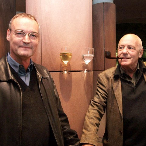Zwei Männer stehen an einer Wand und schauen in die Kamera, zwischen ihnen Weingläser