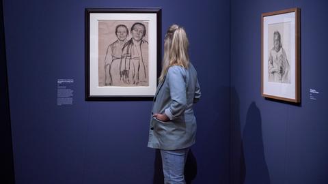 Ausstellungsansicht "Kollwitz" im Frankfurter Städel. Das Bild zeigt eine Frau mit blondem Zopf und hellblauem Hosenanzug. Sie steht in einer Ecke eines dunkelblau gestrichenen Rahmens. An den Wänden hängt jeweils eine gerahmte Kreidezeichnung von Käthe Kollwitz.