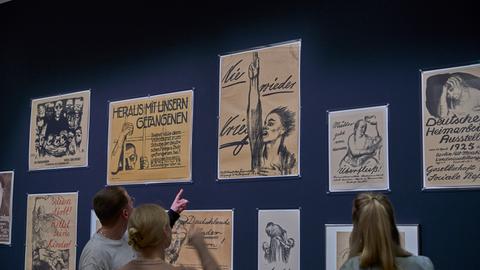 Das Bild zeigt drei Personen, die vor einer blauen Wand stehen. Darauf sind mehrere gezeichnete Plakate angebracht, unter anderem das berühmte Anti-Kriegsplakat "Nie wieder Krieg!" von Käthe Kollwitz.