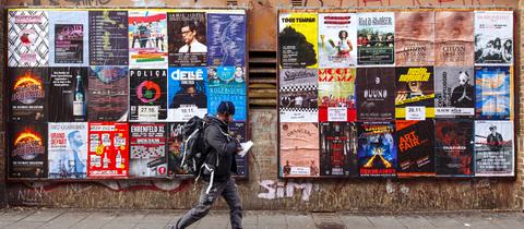 Eine Wand im öffentlichen Raum mit vielen Konzertplakaten. Ein Mann geht vorbei.