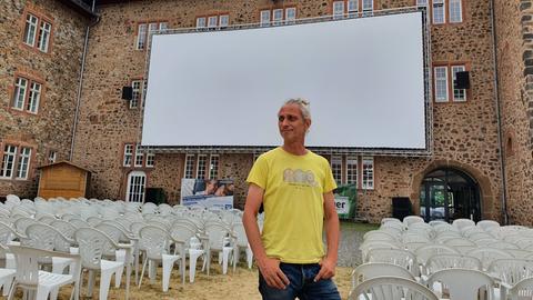 Das Bild zeigt einen Mann mit gelbem T-Shirt und grauem Zopf, der vor einer Open-Air-Kinoleinwand in einem Burginnenhof steht. Hinter dem Mann sind mehrere Reihen weißer Plastikstühle aufgebaut.