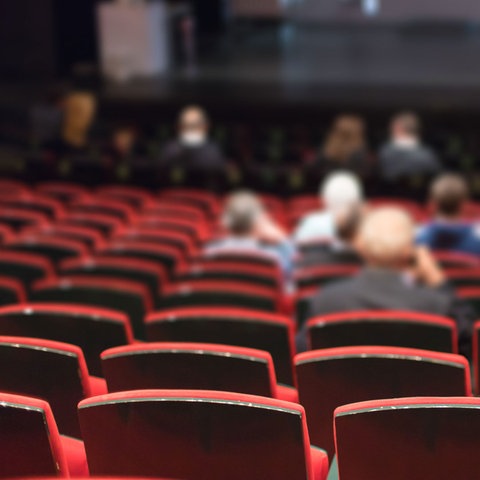 Wenige Menschen sitzen in den Stuhlreihen eines Theaters. Viele Plätze bleiben leer. 