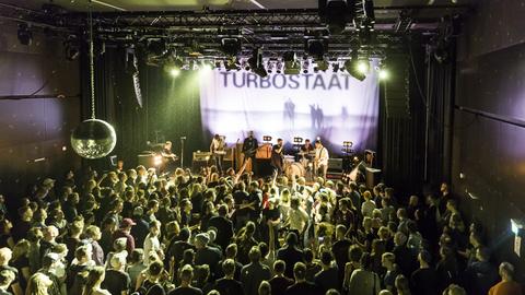 Das Bild zeigt aus der Vogelperspektive einen gut gefüllten Konzertraum und eine Band auf der Bühne. Auf einem Bildschirm leuchtet das Wort "Turbostaat".