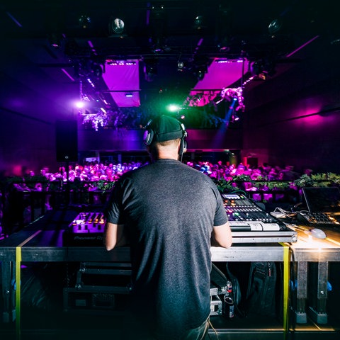 Das Bild zeigt eine Party in einem lila beleuchteten Club. Ein DJ steht mit dem Rücken zur Kamera an seinem Pult. 