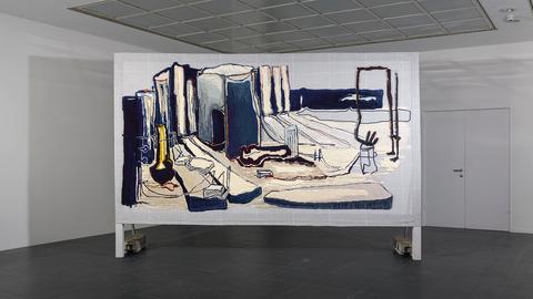 Großformatiger Wandteppich in Blau- und Grautönen auf einer Stellfläche in einem Ausstellungsraum.