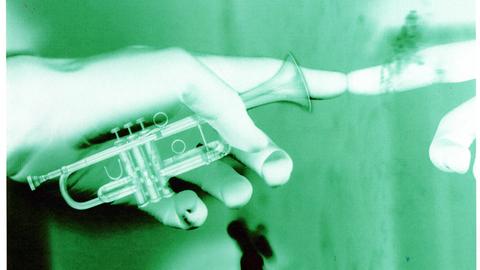 Das Bild zeigt eine Hand mit ausgestrecktem Zeigefinger, der auf einen Spiegel zeigt. So scheint er sich selbst zu berühren. Die Hand hält außerdem eine kleine Tuba fest.