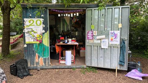 Das Bild zeigt einen Container auf einem mit Rindenmulch ausgelegten Platz. Er besteht aus Metall. Die offenen Türen sind mit von Kindern gemalten Bildern beklebt. Im Inneren sind ein Werktisch und Bastelmaterialen zu sehen.