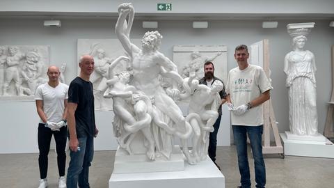 Vier Männer stehen um eine antike Figurengruppe in der Optik von weißem Marmor