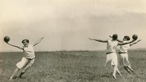 Das Bild zeigt drei Frauen im Jahr 1930, die in weißen Sportanzügen Gymnastik mit Ball auf einer Wiese betreiben