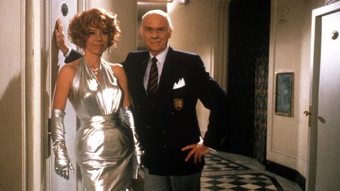 Eine Frau in silbernem Kleid und ein Mann in einem Hotel-Flur.