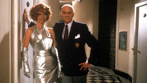 Eine Frau in silbernem Kleid und ein Mann in einem Hotel-Flur.