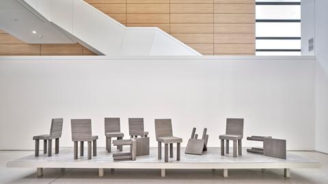 9 Holzstühle stehen auf einem weißen Podest in einer Halle mit hoher Decke und einer Fassade aus Holz und Glas.