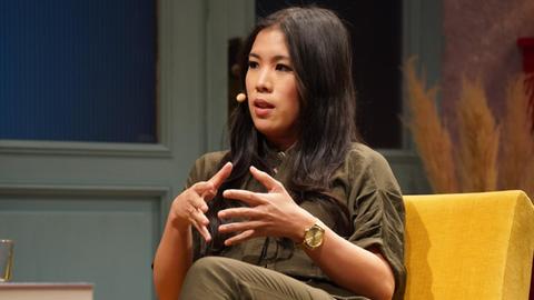 Mai Thi Nguyen-Kim sitzt auf einem gelben Sofa. Sie trägt einen khakifarbenen Overall und ihre dunklen Haare fallen über ihre Schulter.