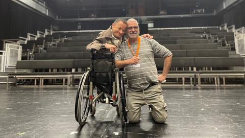 Zwei Männer, einer sitzt im Rollstuhl, auf einer Theaterbühne.