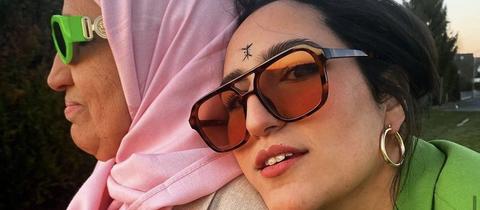 Eine junge Frau mit Sonnenbrille neben einer älteren Frau mit rosa Kopftuch.